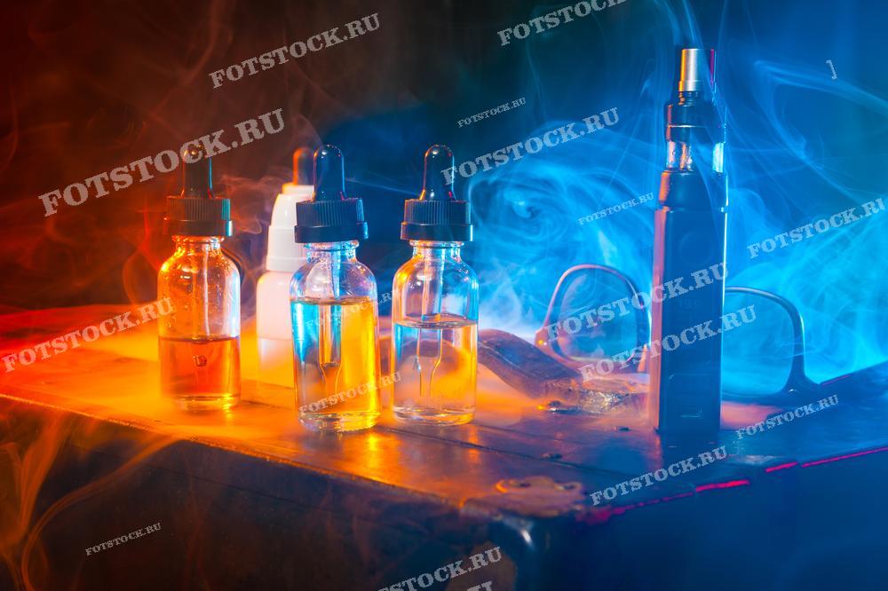 На фотографии изображены несколько флаконов с жидкостью для вейпинга и вейп-устройство, стоящие на деревянном столе.